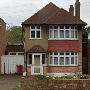 A two-story residential property with a beige exterior, red roof tiles, and white windows, situated in a garden area with a driveway.